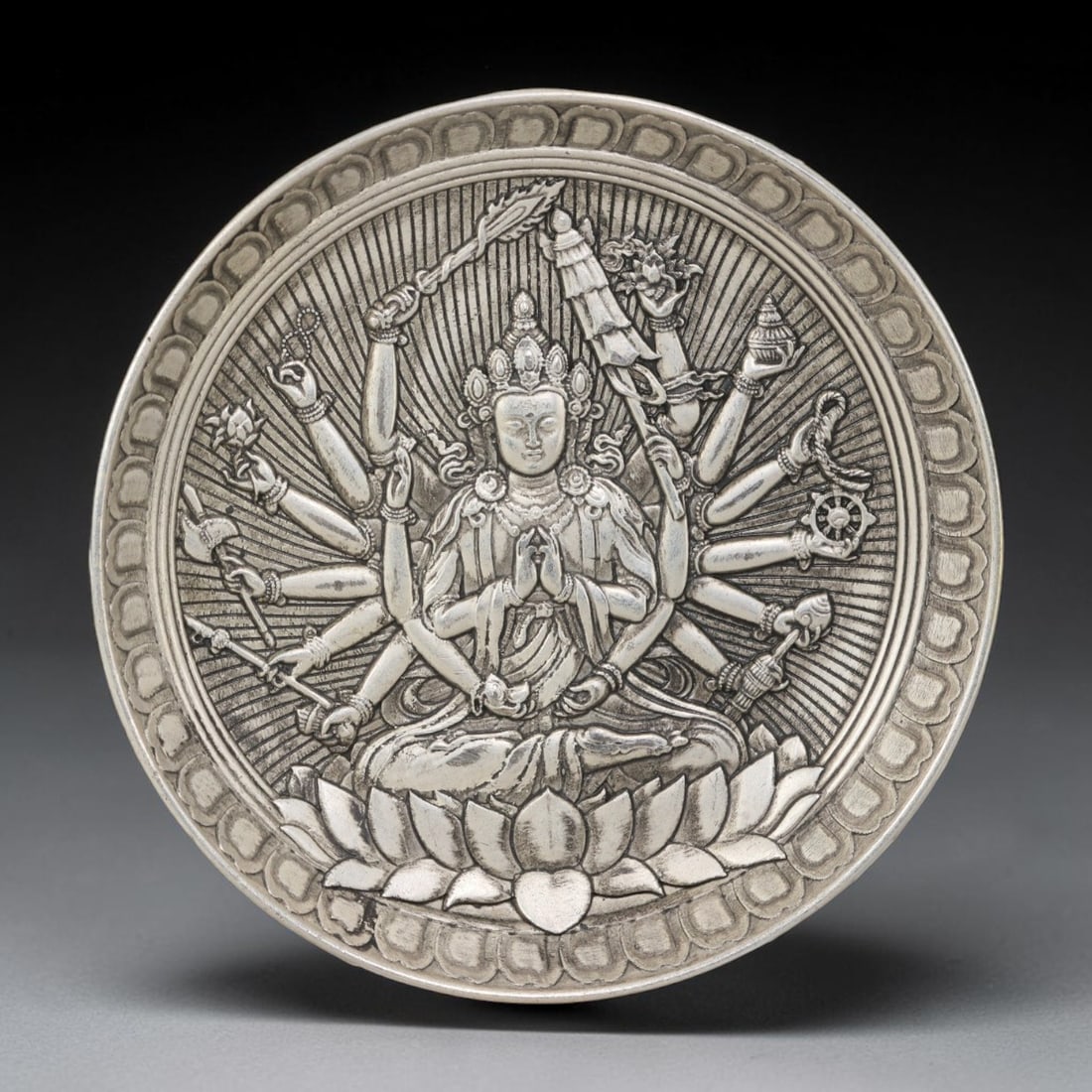 A Tibetan Thousand-Armed Avalokiteshvara German Silver Decorative Plate, 28AD: A circular plate in German silver (a nickel-silver alloy), the center decorated in high relief with a thousand-armed Avalokiteshvara. The deity is shown seated in a meditative posture upon a lotus thr