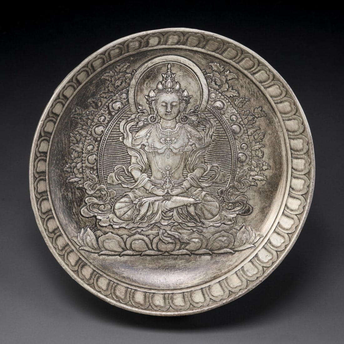 A Seated Deity German Silver Decorative Plate, 30AD: A circular plate in German silver (a nickel-silver alloy), the center decorated in high relief with a seated Buddhist figure. The figure is shown in a meditative posture upon a lotus throne, wearing a