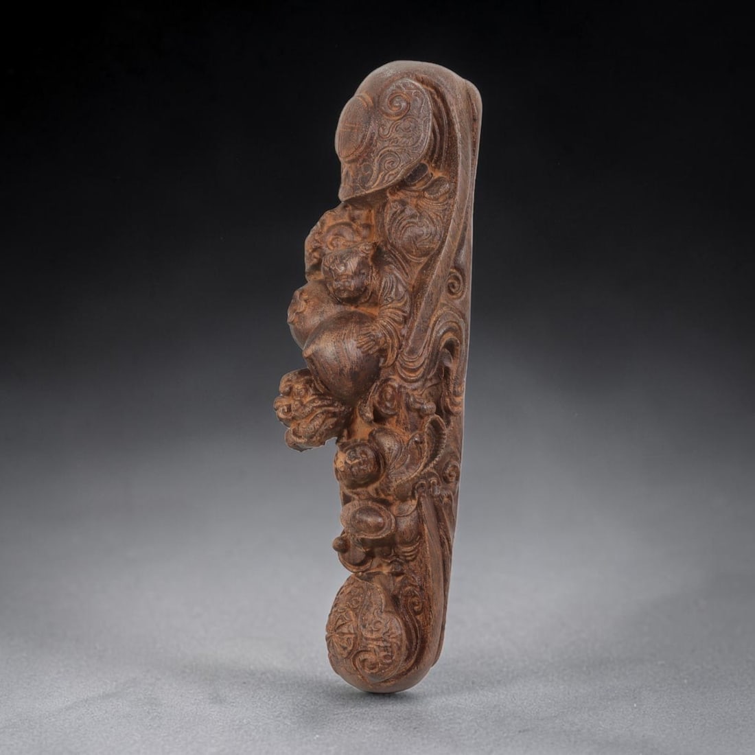 A Carved Agarwood Ruyi Scepter Sculpture, 8AD: Carved from fragrant agarwood (Aquilaria spp.), the work takes the form of an elongated Ruyi scepter with a cloud-head terminal. The shaft is decorated in high relief with figures of children, mythica