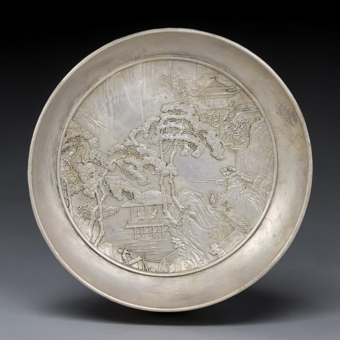 A Chinese Landscape German Silver Decorative Plate, 26AD: A circular plate in German silver (a nickel-silver alloy), the center decorated in high relief with a Chinese landscape composition. The scene depicts ancient pine trees, layered rock formations, flow