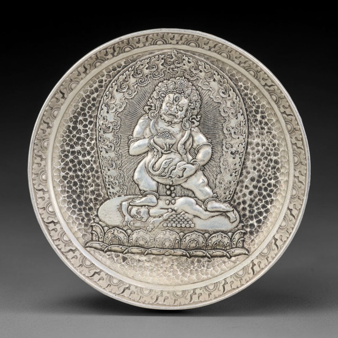 A Tibetan Black Jambhala German Silver Decorative Plate, 33AD: A circular plate in German silver (a nickel-silver alloy), the center decorated in high relief with a Black Jambhala figure in a Tibetan Buddhist manner. The deity is shown standing upon a lotus base,