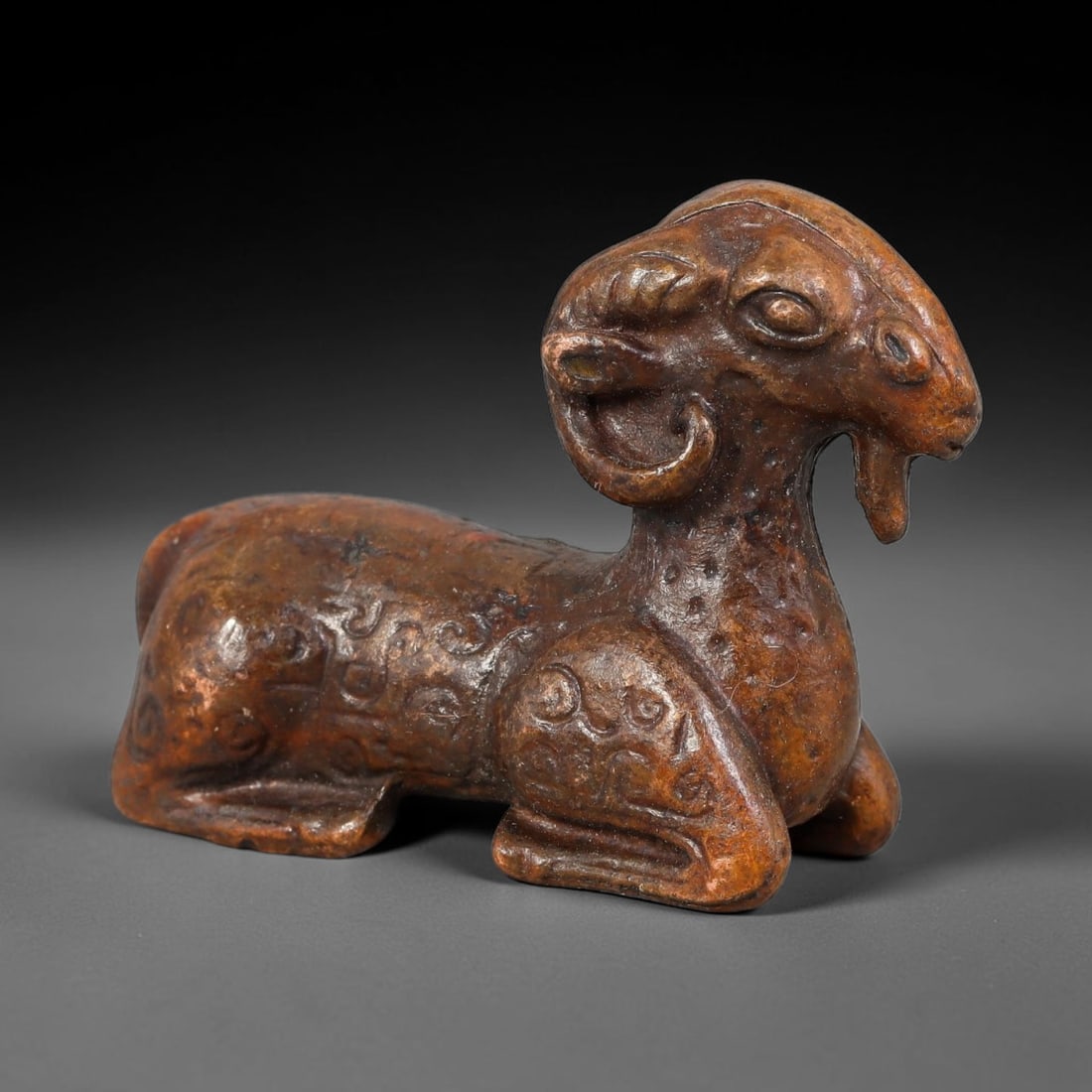 A Chinese Bronze Recumbent Ram Figurine Effigy, 19AD: Cast in bronze, this figurine depicts a ram in reclining pose with prominent curved horns, stylized facial features and hanging tongue detail. The body, neck and limbs are adorned with deeply-cast arc