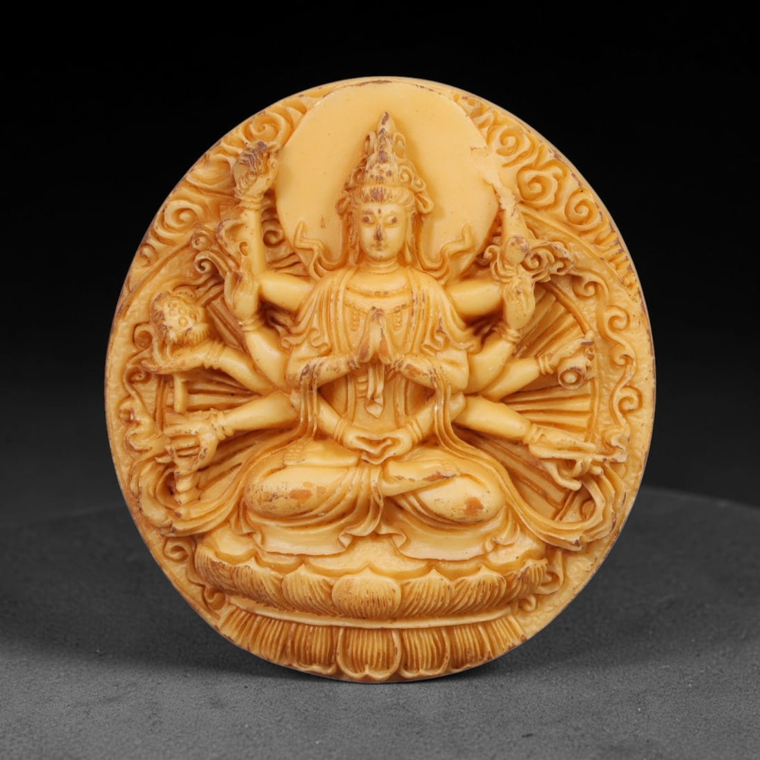 A Chinese Multi-Armed Guanyin Deity Carved Tagua Nut Pendant Amulet, 85BC: Carved from tagua nut, this circular tablet-form amulet is executed in high relief. The multi-armed Guanyin sits in lotus position upon a detailed lotus pedestal, central hands pressed together in anj