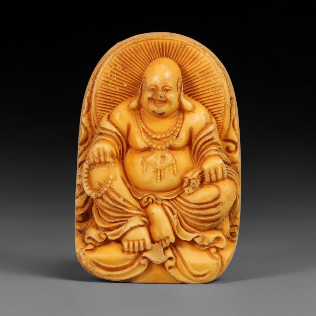 A Chinese Laughing Buddha Carved Tagua Nut Pendant Amulet, 93BC: Carved from tagua nut, this rounded oval tablet-form amulet features high-relief carving of the laughing Budai Buddha. The figure is depicted in relaxed seated pose with broad joyful expression, full 