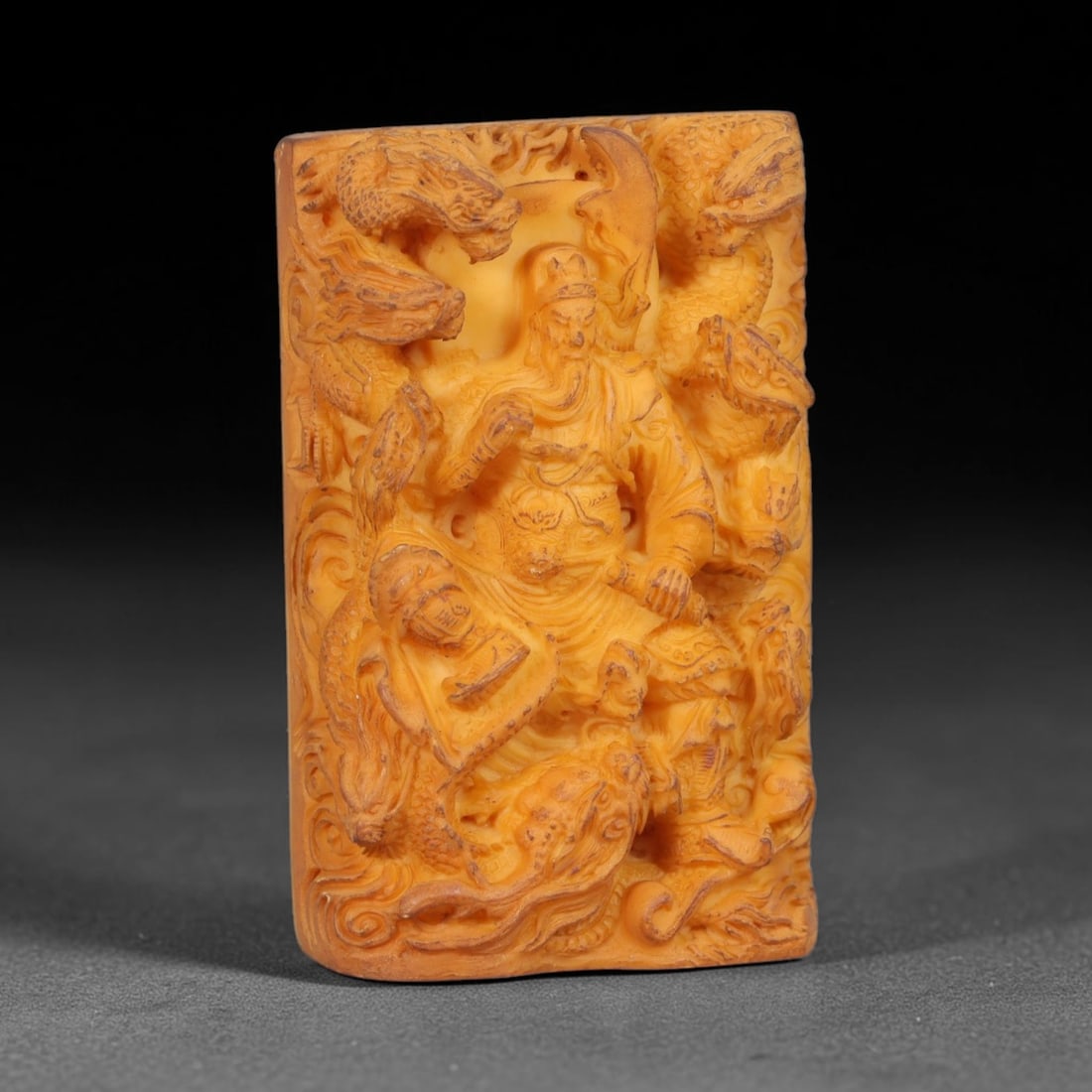 A Chinese Guan Gong Deity Carved Tagua Nut Pendant Amulet, 5AD: Carved from tagua nut, this rectangular tablet-form amulet is rendered in high relief. The central figure depicts Guan Gong, shown in traditional warrior regalia with defined facial hair and headdress