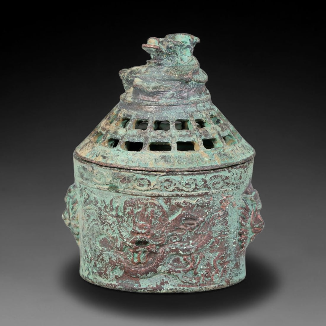 A Chinese Bronze Dragon-Finial Lidded Censer, 57AD: Cast in bronze, this compact lidded censer has a cylindrical body decorated in relief with a coiled dragon motif flanked by stylized mask handles. The domed lid features lattice openwork perforations 