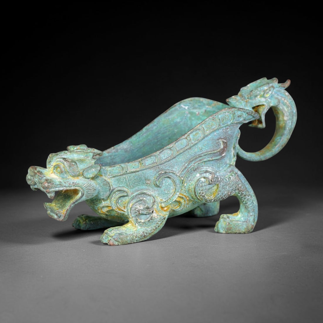 A Chinese Archaic-Style Bronze Mythical Beast Pouring Vessel with Dragon-Head Handle, 19AJ: Cast bronze trough-form pouring vessel, modelled as a striding mythical beast with an open-mouthed beast-head spout at the front, supported by four detailed clawed legs. A looping dragon-head forms th