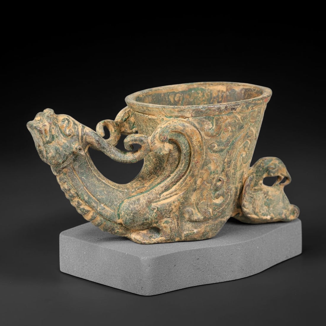 A Chinese Archaic-Style Bronze Dragon-Form Rhyton Vessel with Phoenix Terminal, 56AD: Cast bronze rhyton-style libation vessel featuring a prominent dragon-head spout forming the forward base, with scrolling tendril attachments connecting to the tapered cup body. A recumbent open-work 