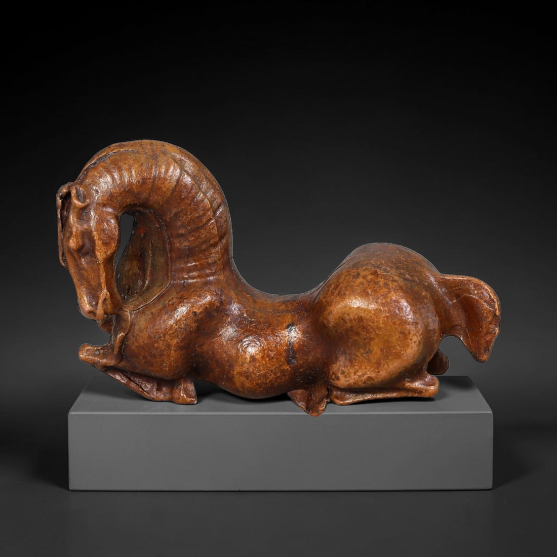 A Chinese Bronze Recumbent Curled-Neck Horse Figurine, 50AD: Cast bronze figurine depicting a recumbent horse with elegantly arched, looping neck and dynamically curled head, finely modelled musculature and mane details. Warm mellow amber-brown aged patina cove