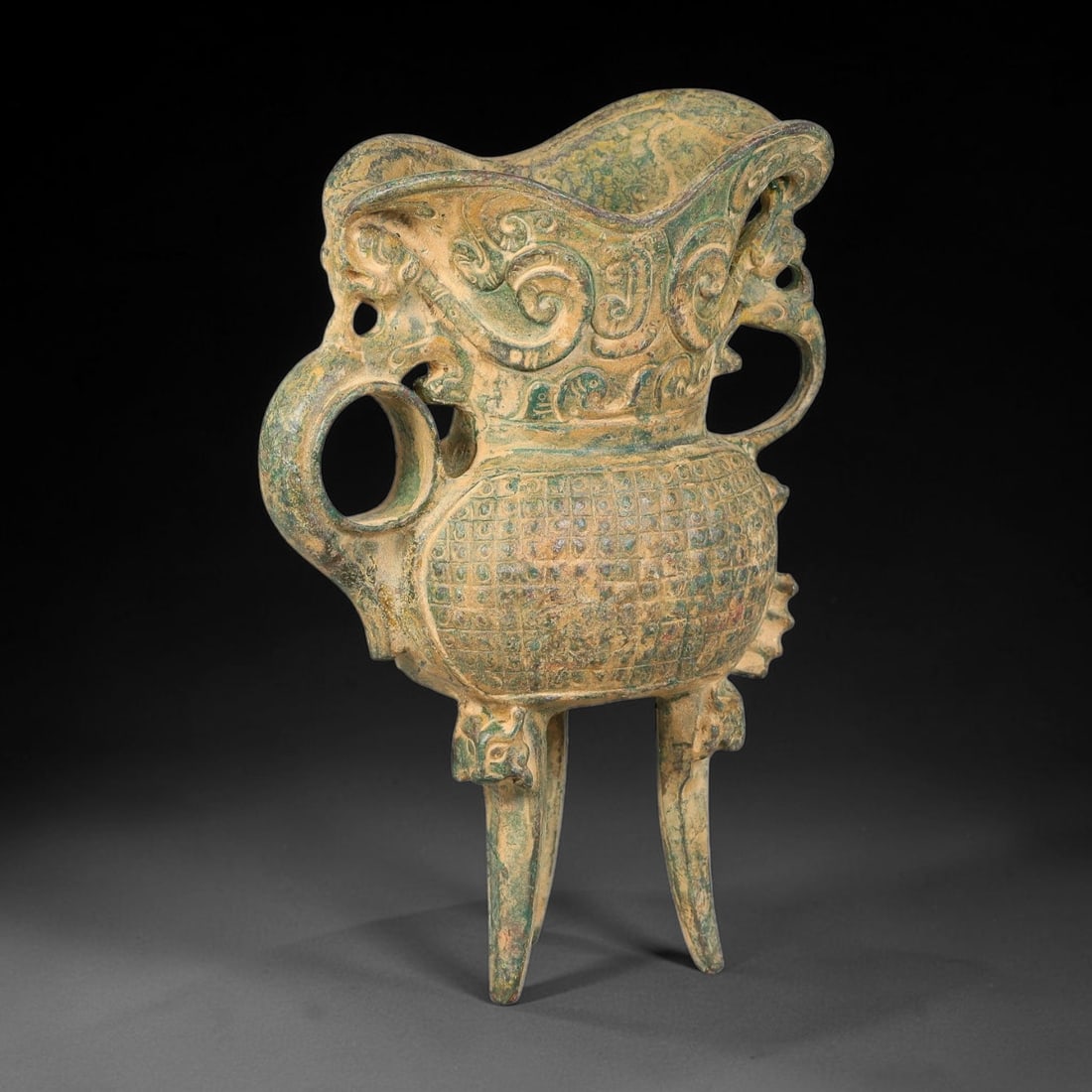 A Chinese Archaic-Style Bronze Tripod Ritual Wine Vessel, 80AD: Cast bronze tripod ritual wine vessel with wide scalloped flared rim, flanked by two open-work beast-form side handles. Upper section decorated with flowing scroll-pattern relief, main body covered in