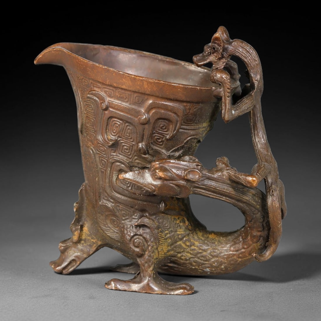 A Chinese Archaic-Style Bronze Dragon-Handled Libation Cup, 88AD: Cast bronze libation cup with curved pouring spout, the body deeply cast with archaic spiral-scroll relief patterns. It is supported by a stylized dragon-form base and fitted with an open-work dragon-