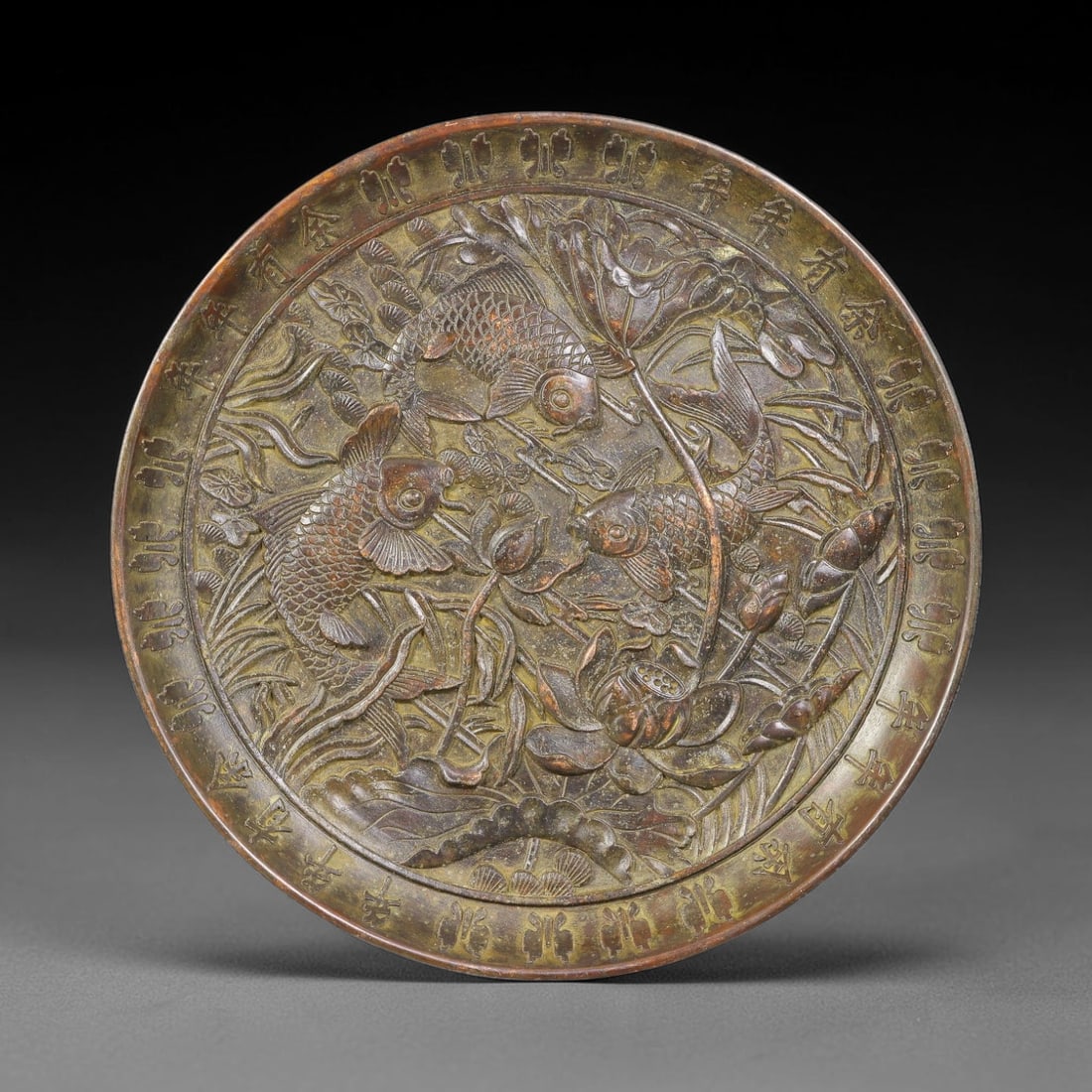 A Chinese Bronze Relief Plate with Carp and Lotus Motif, 21AJ: Cast in bronze, this circular plate features high-relief decoration depicting three carp swimming amid full-bloom lotus blossoms, buds and aquatic foliage. The surrounding rim is inscribed with repeat