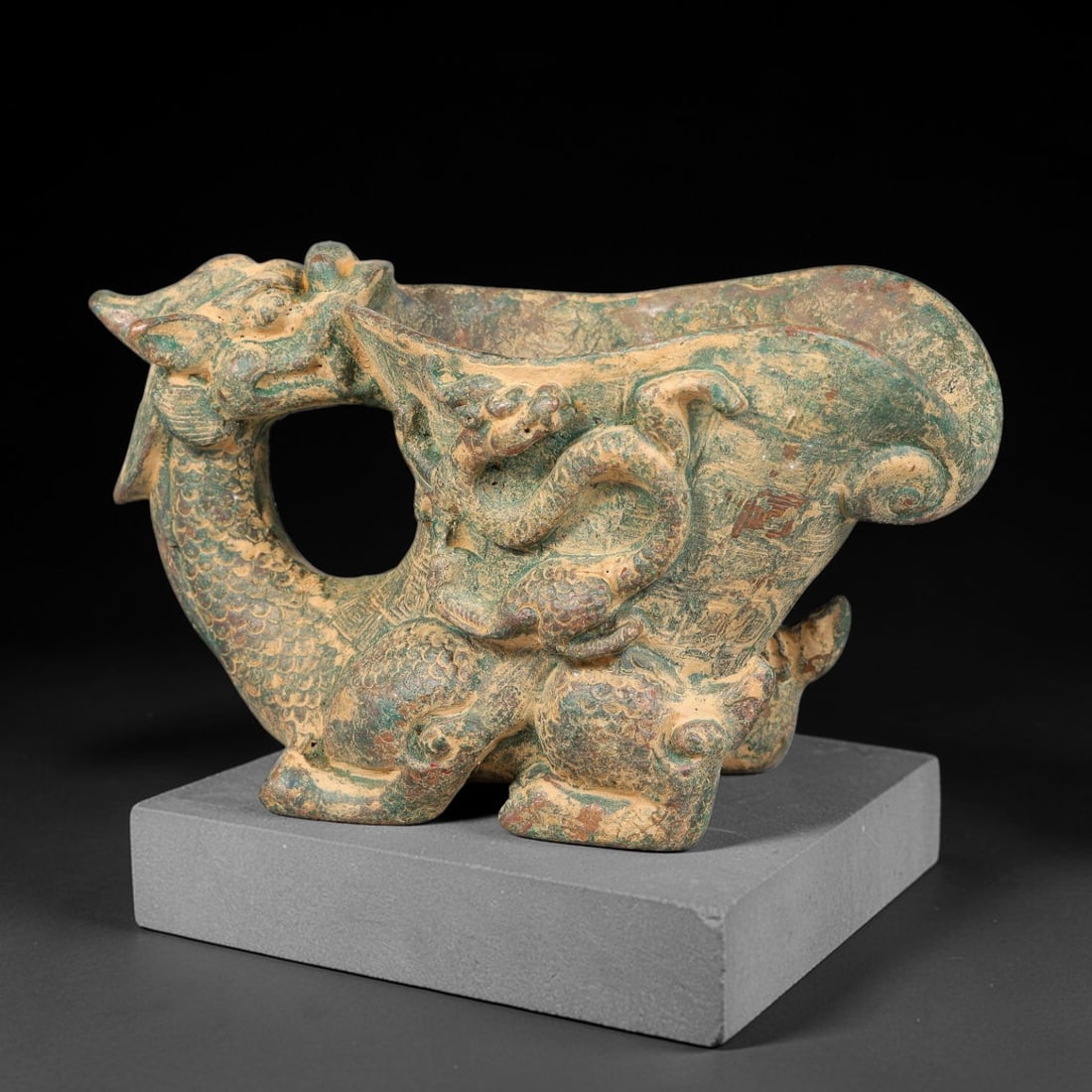 A Chinese Archaic-Style Bronze Dragon-Body Libation Vessel, 83AD: Cast bronze libation vessel supported by a crouching scaled dragon figure whose arched neck and head forms the integrated looping handle. The wide flared trough-shaped body is adorned with raised arch