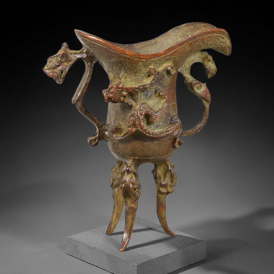 A Chinese Archaic-Style Bronze Dragon-Ornamented Jue Wine Vessel, 9AJ: Cast bronze ritual jue wine vessel with wide flared spout and elegant profile, fitted with two stylized beast-form side handles. A relief coiled dragon adorns the front body, supported by three ornate