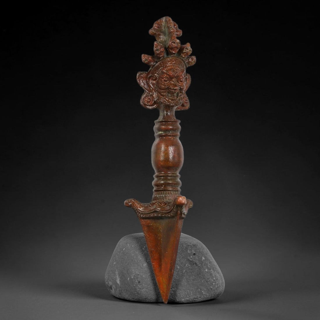 A Chinese Tibetan Bronze Phurba Ritual Dagger, 22AD: Cast in bronze, this ceremonial phurba dagger is surmounted by a detailed wrathful deity mask surrounded by stylized foliate ornament. The turned bulbous handle features moulded ringed bands, descendi