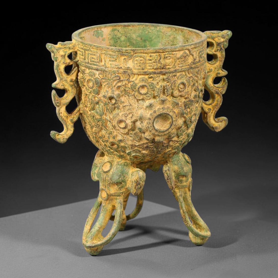 A Chinese Archaic-Style Bronze Tripod Vessel with Dragon Handles and Elephant Legs, 79AD: Cast bronze tripod vessel with two elaborate open-work dragon-form side handles. The rounded bowl body is densely covered in high-relief scrolling floral motifs, topped with an engraved key-fret borde