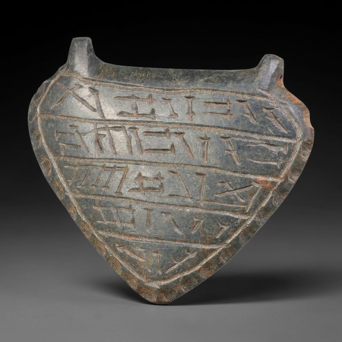 Ancient Uruk Period Hard-Stone Heart-Shaped Inscribed Apotropaic Amulet (c.3500-3100 BCE), 66AJ: Heart-form apotropaic amulet originating from the Uruk cultural sphere, featuring multiple horizontal registers of incised archaic symbolic script running across its surface. Two suspension lugs are i