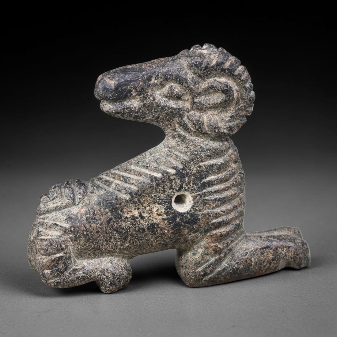 Ancient Sasanian Persian Schist Crouching Ram Talisman Amulet (224-651 CE), 58AJ: Dark schist zoomorphic amulet formed as a recumbent ram, a prominent apotropaic motif within Sasanian Persian folk ritual craft. Incised linear detailing defines the curled horn structure, mane and bo