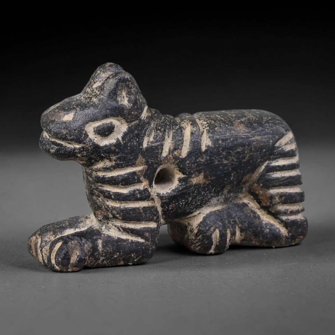 Ancient Elamite Civilization Recumbent Feline Stone Amulet (3200-539 BCE), 72AJ: This carved dark stone recumbent feline amulet originates from Elamite Civilization, detailed with incised linear patterning across body, limbs and tail. A central perforation through the torso allows