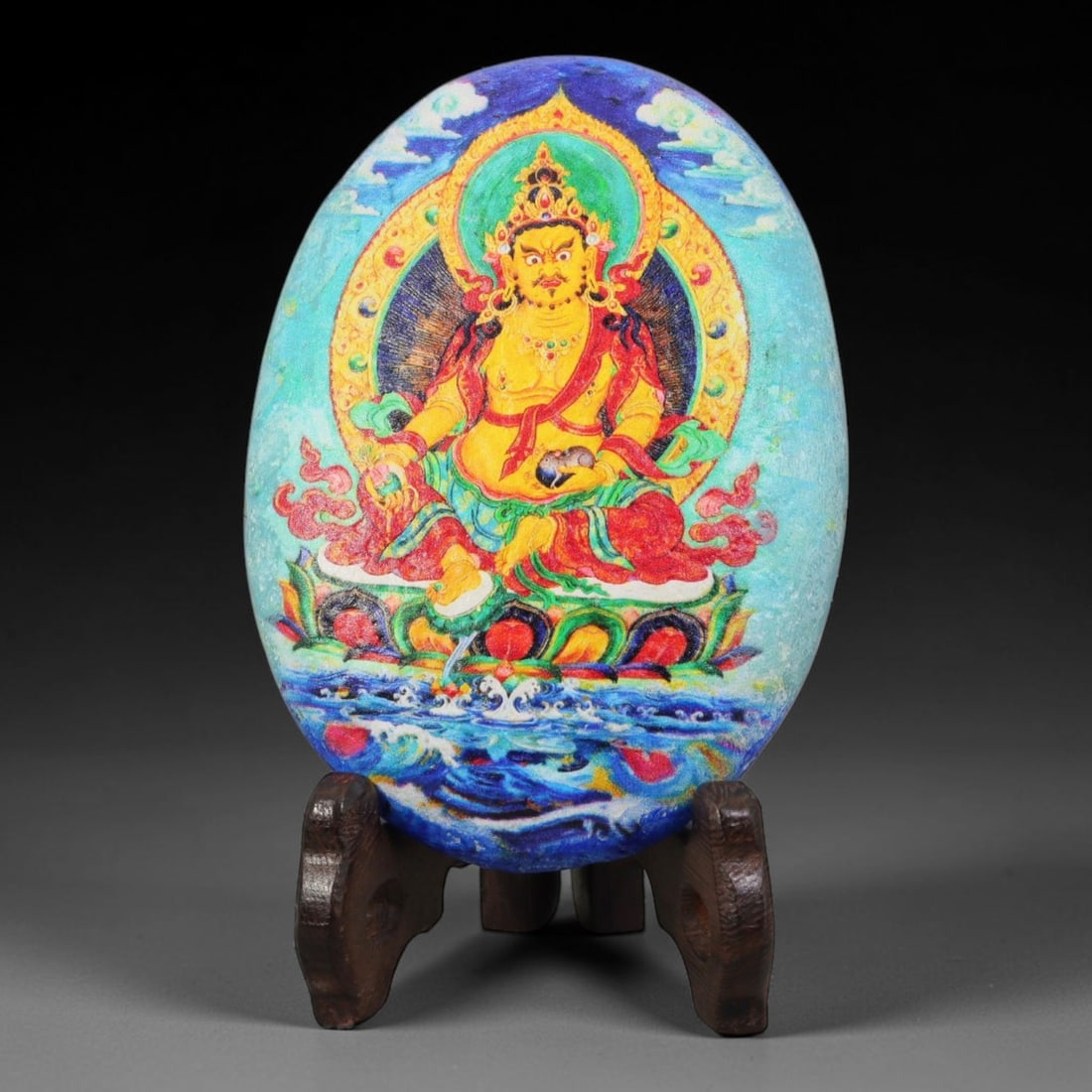 A Tibetan Thangka Style Painted River Stone with a Wealth Deity, 77BC: Painted on a naturally rounded river stone, the work depicts Yellow Jambhala, a wealth deity, in a thangka-style composition. The figure is shown with a golden body, wearing red-and-green robes and se