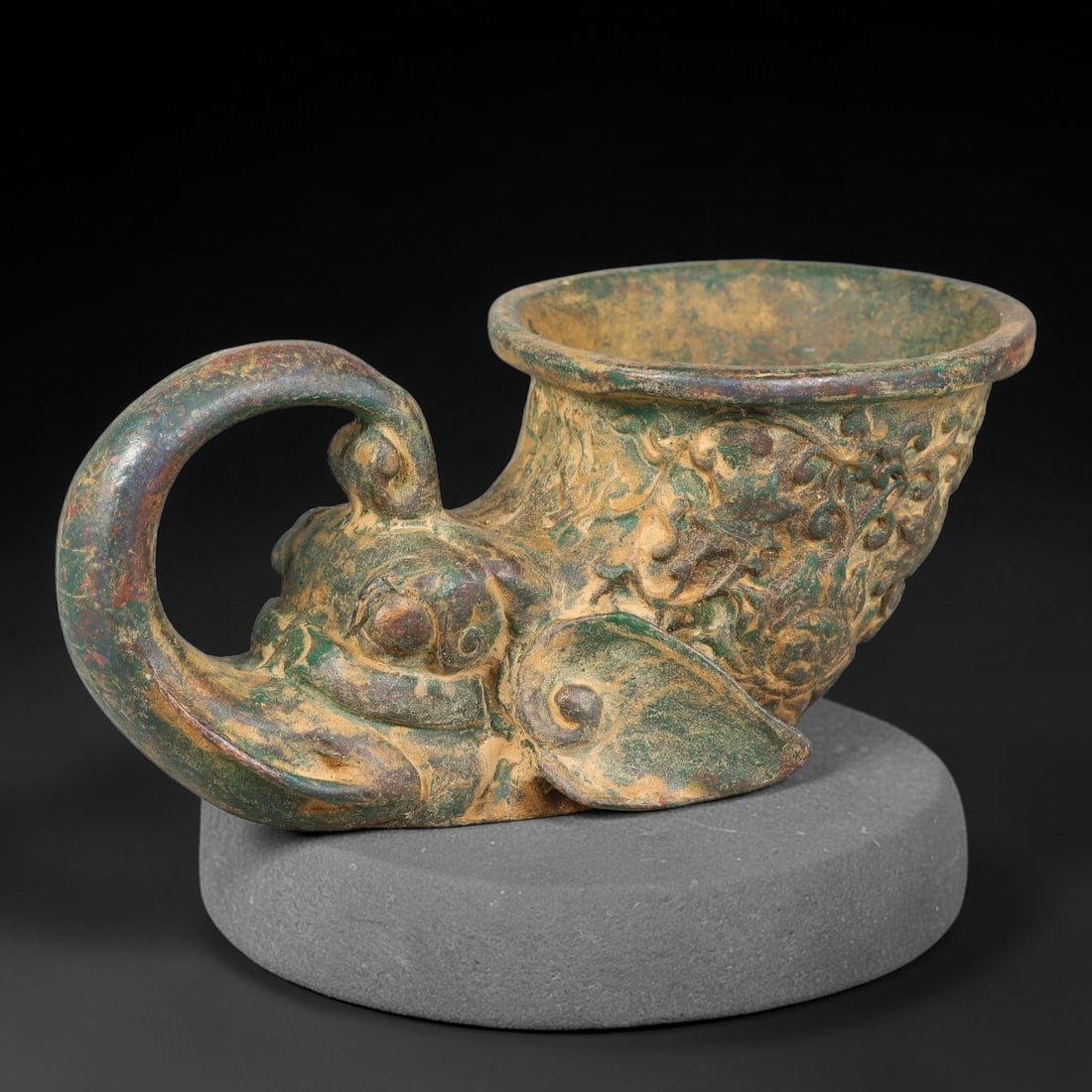 A Chinese Archaic-Style Bronze Elephant-Head Rhyton Vessel, 81AD: Cast bronze horn-shaped rhyton libation cup anchored by a finely modelled elephant-head forming the base and looping curved handle. The tapered cup body is adorned with scrolling floral and foliate re