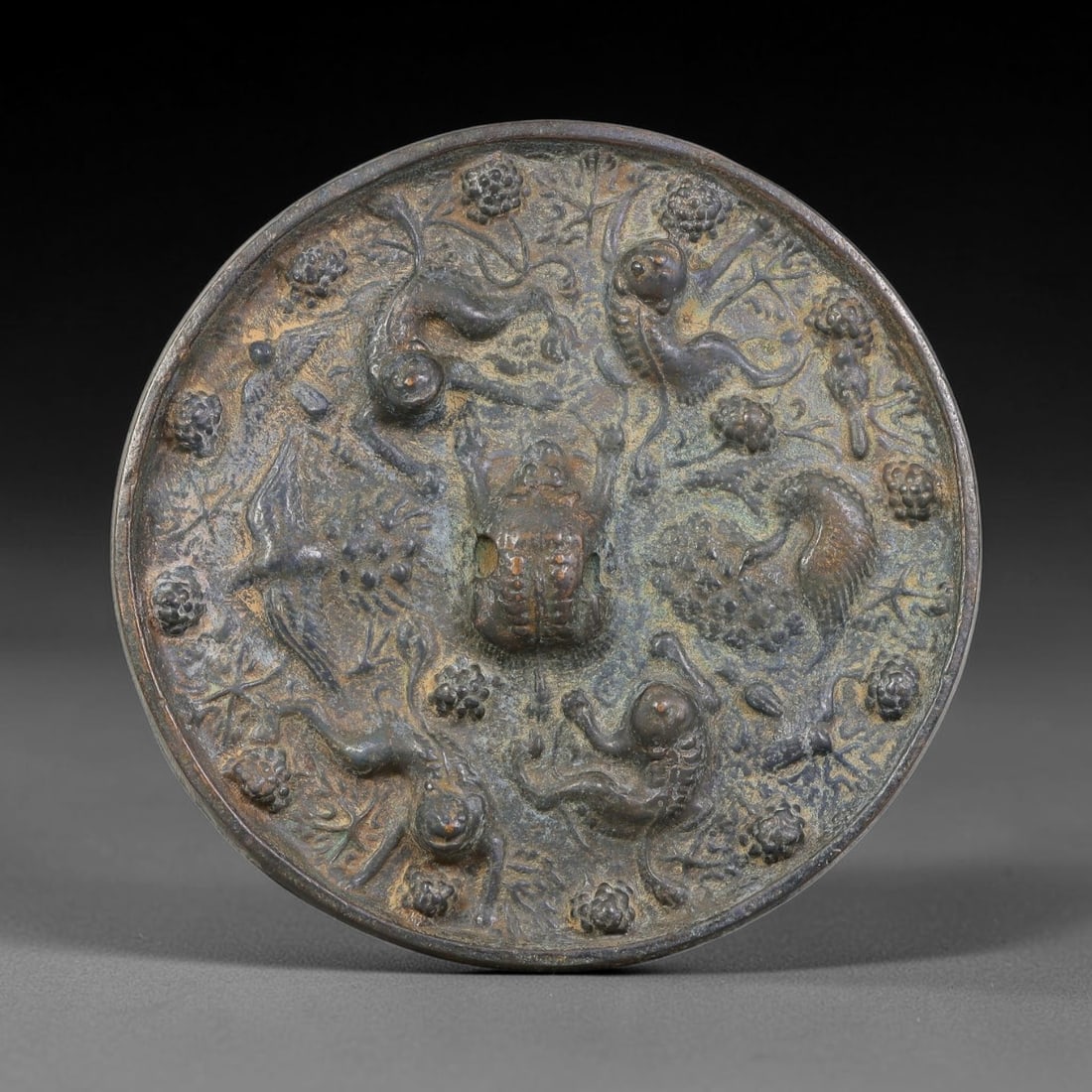 A Chinese Archaic-Style Bronze Mirror with Beasts Birds and Grapevine Relief, 25AJ: Cast bronze circular mirror featuring a central tortoise-form knob, its face densely decorated in high relief with roaming mythical beasts, birds and scrolling grape-vine foliate motifs covering the e