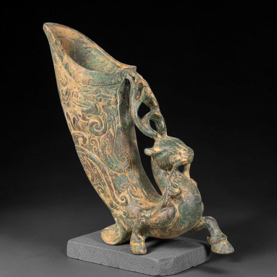 A Chinese Archaic-Style Bronze Deer-Supported Rhyton Libation Vessel, 82AD: Cast bronze horn-shaped rhyton libation cup upheld by a dynamically modelled striding deer forming the handle and base, with open-work antler-form looping connections to the vessel body. The horn-form
