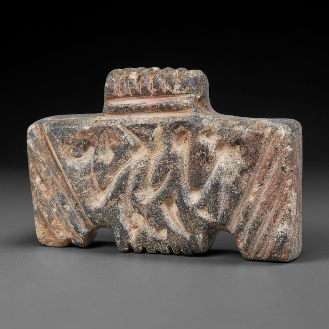 Ancient Elamite Civilization Relief-Carved Figural Stone Amulet (3200-539 BCE), 87AJ: This rectangular carved dark stone amulet originates from Elamite Civilization, displaying stylized relief figural motifs on its main face, framed by incised linear border ornamentation. A notched upp