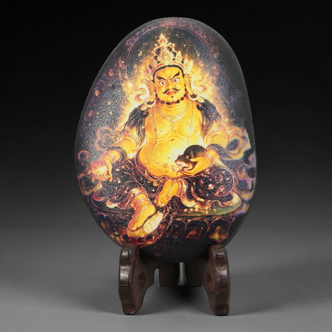 A Tibetan Thangka Style Painted River Stone with a Wealth Deity, 73BC: Painted on a naturally rounded, oval black river stone, the work depicts Yellow Jambhala, a wealth deity, in a thangka-style composition. The figure is shown with a golden body and an imposing counten