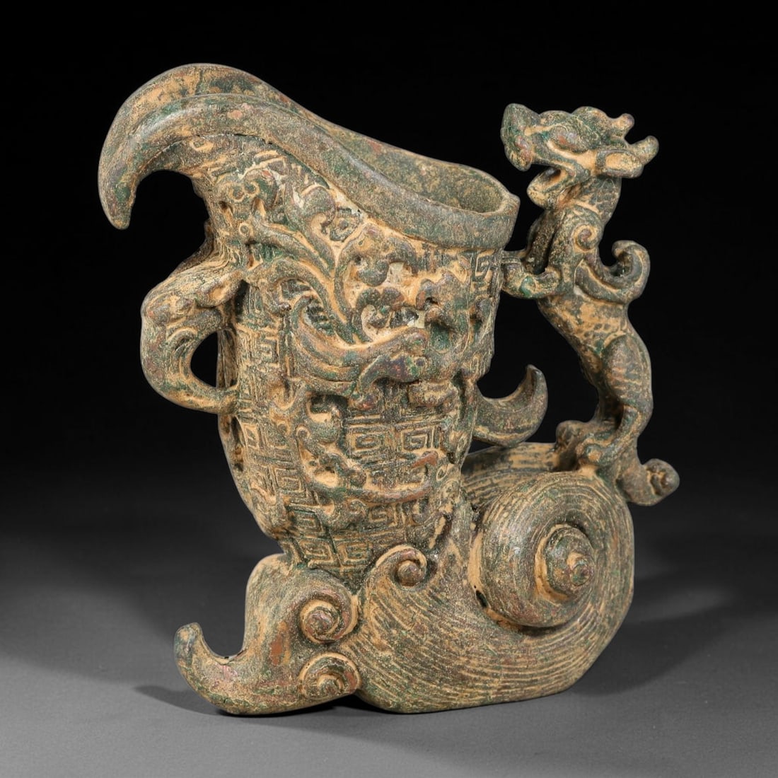 A Chinese Archaic-Style Bronze Rhyton Vessel with Dragon Handle and Scroll Base, 64AD: Cast bronze rhyton libation vessel with upward-curving flared spout, supported by a large coiled scrolling cloud-form base. A fully modelled standing dragon forms the prominent side handle, while a sm