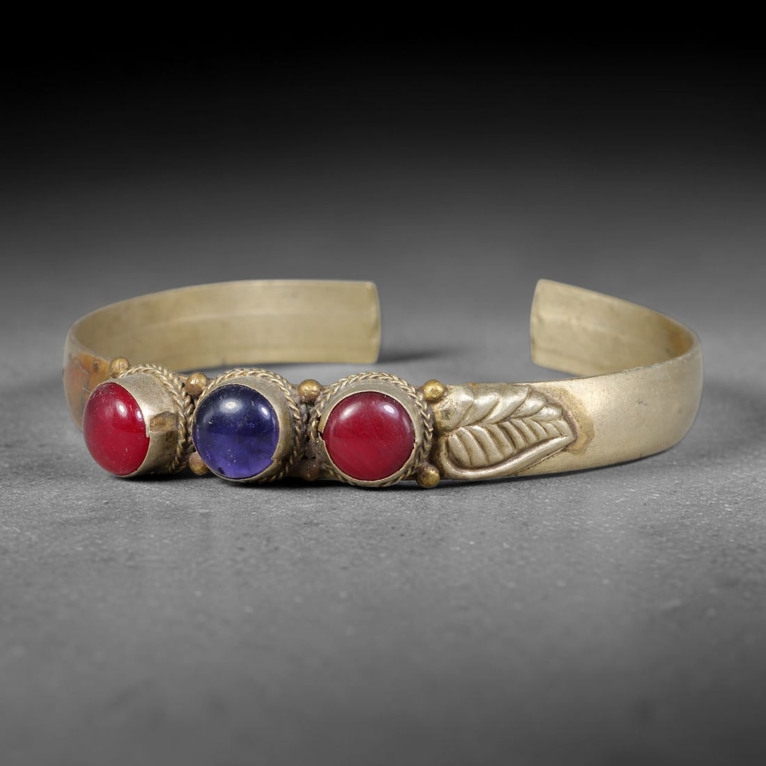 A Central Asian Copper-Alloy Multi-Stone Inlaid Open-Cuff Bracelet, 52AK: Open-form copper-alloy cuff bracelet set with three round cabochon insets held within twisted wire bezels, accompanied by small spherical metal studs between each setting. A single repousse leaf-shape