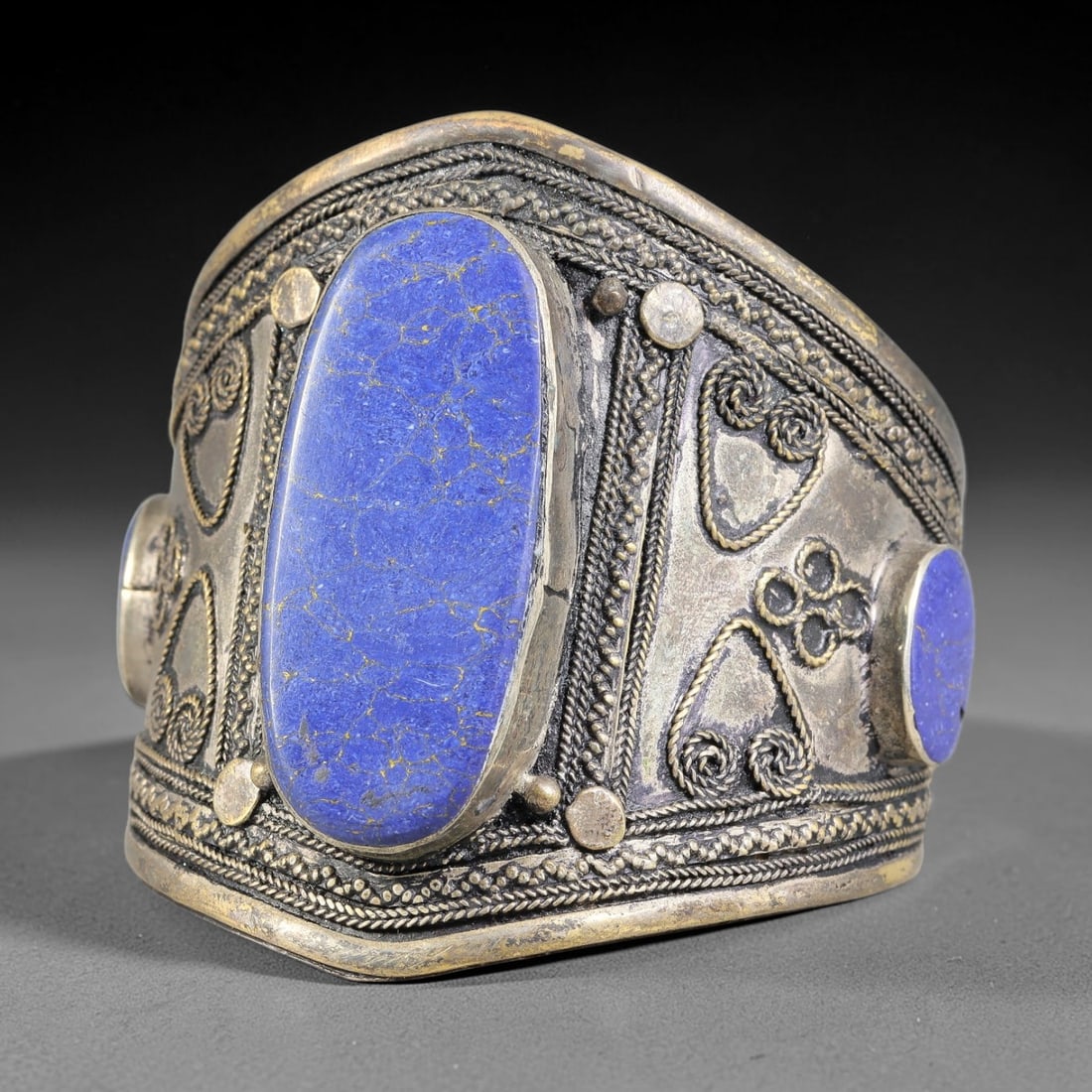 A Central Asian Copper-Alloy Lapis Lazuli Inlaid Cuff Bracelet, 38AK: Stout copper-alloy cuff bracelet centering a large elongated lapis lazuli cabochon within bezel mounting, flanked by secondary round inlaid stones. The surface is ornamented with applied twisted filig