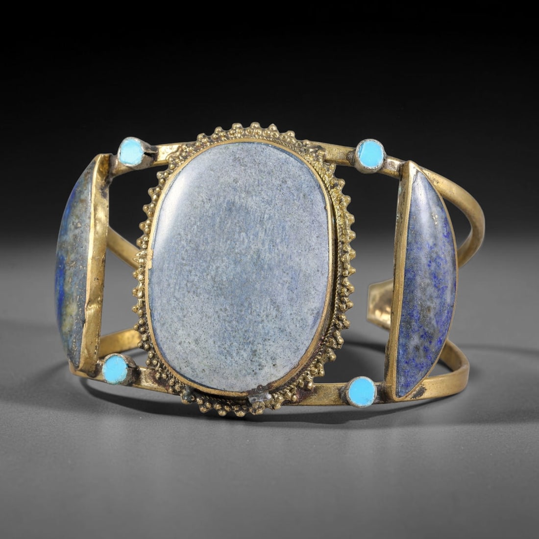 A Central Asian Gilt-Metal Lapis Lazuli and Turquoise Open Cuff Bracelet, 42AK: Open-form gilt-metal cuff bracelet centering a large oval cabochon, bordered by granulated beading. Elongated lapis lazuli elements flank the central stone, accented by small round turquoise inlays se