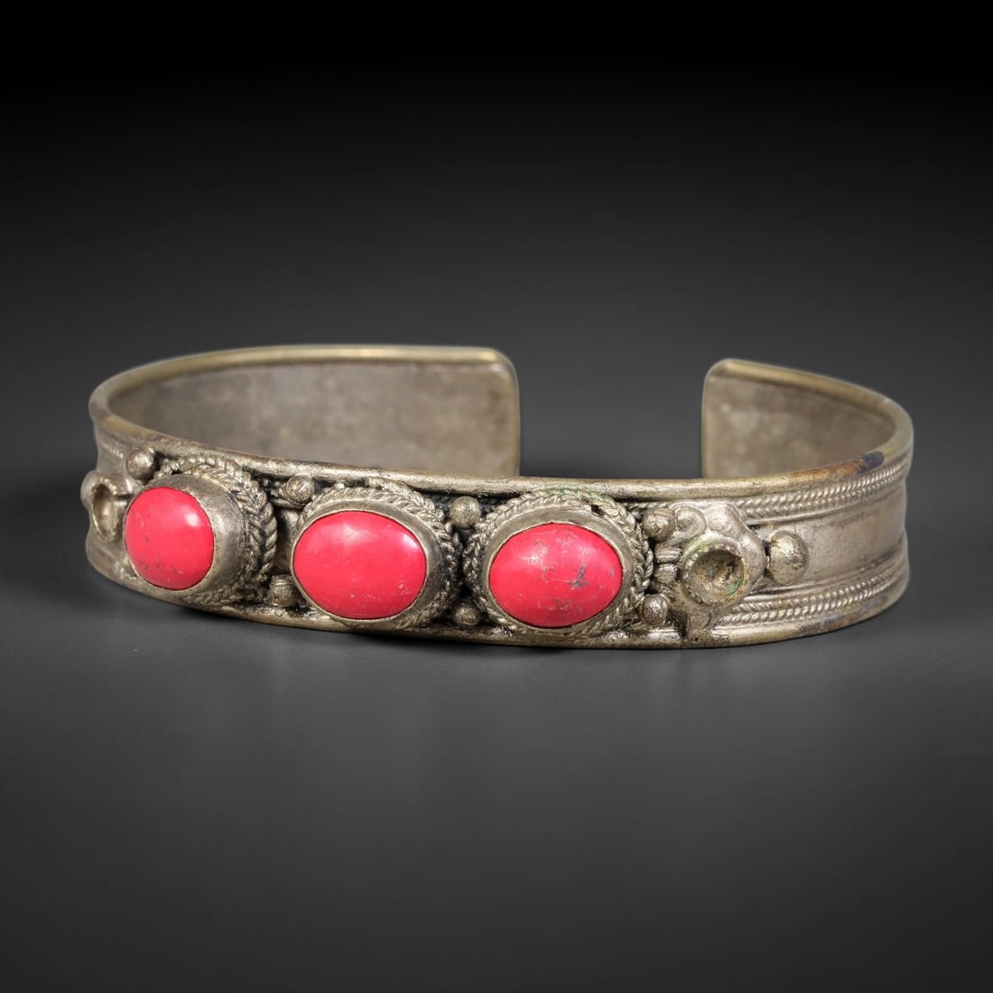 A Central Asian Copper-Alloy Red Composite Inlaid Open-Cuff Bracelet, 51AK: Open-cut copper-alloy cuff bracelet displaying three oval red composite cabochons each nested within twisted wire bezel surrounds. Raised spherical studs and multi-strand incised wirework decorate the