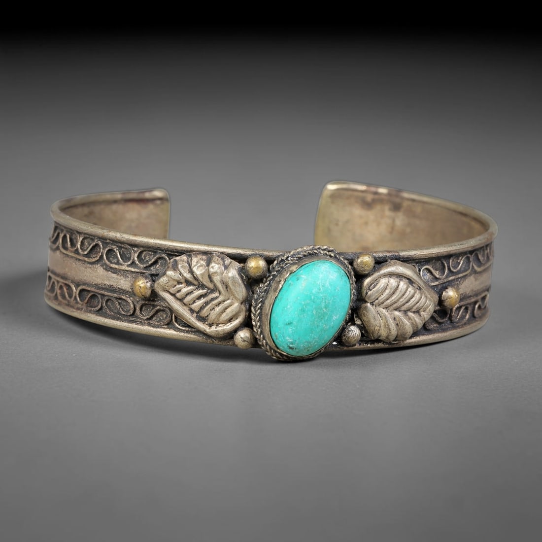 A Central Asian Copper-Alloy Turquoise Inlaid Open-Cuff Bracelet, 50AK: Open-form copper-alloy cuff bracelet set with a central oval turquoise cabochon secured within a twisted-wire bezel. Repousse leaf-shaped relief elements flank the stone, accompanied by raised spheric