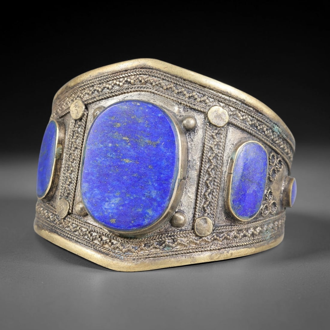 A Central Asian Copper-Alloy Lapis Lazuli Inlay Cuff Bracelet, 44AK: Heavy gauge copper-alloy cuff bracelet fitted with multiple oval lapis lazuli cabochons set within bezels. The band is decorated with applied twisted wire filigree and raised stud ornamentation framin