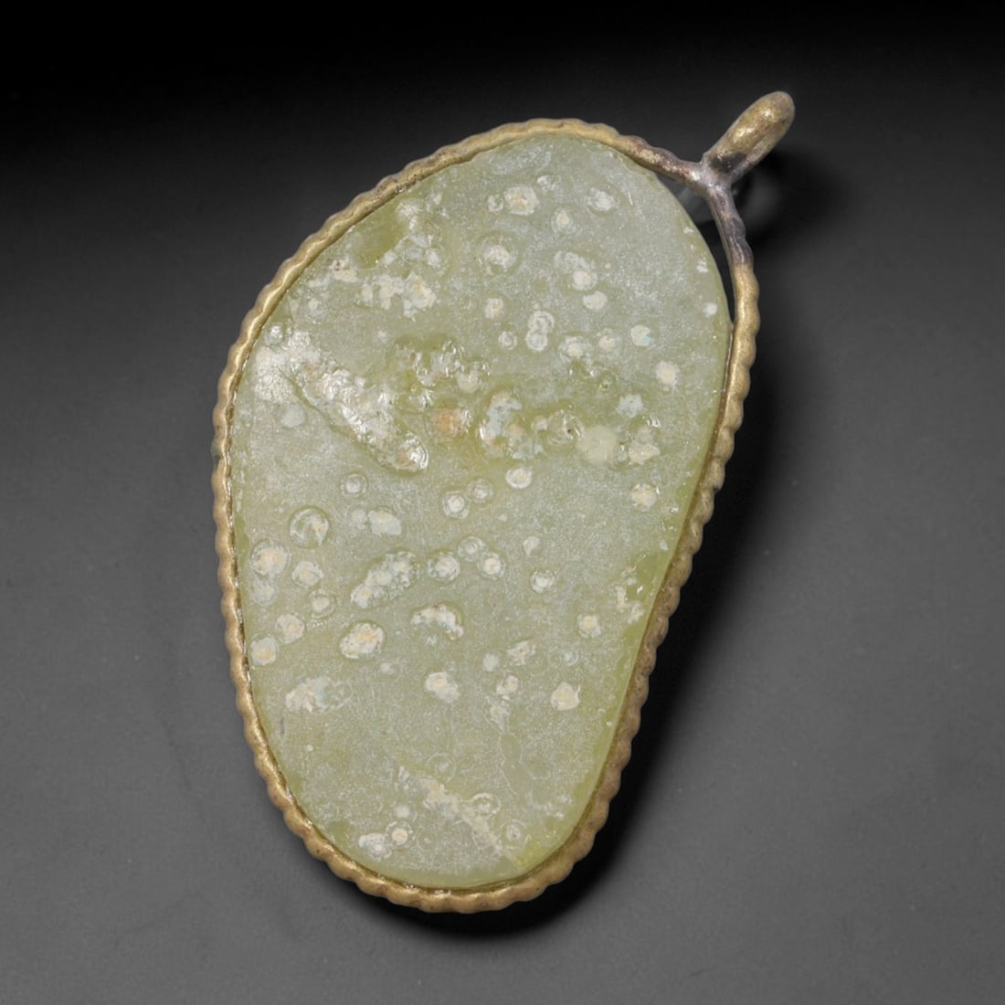 Ancient Roman Glass Slag in Modern Gilt-Edge Pendant, 75AK: This contemporary pendant is crafted from ancient Roman glass slag, featuring a fragmented, iridescent glass surface with weathered patina. The irregular form is bordered by a subtle gilt frame, added
