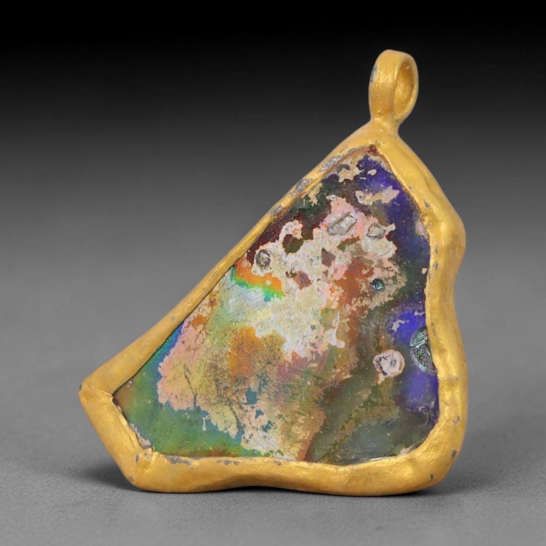 Ancient Roman Glass Slag in Modern Gilt-Edge Pendant, 60AK: This contemporary pendant is crafted from ancient Roman glass slag, featuring a fragmented, iridescent glass surface with weathered patina. The irregular form is bordered by a subtle gilt frame, added