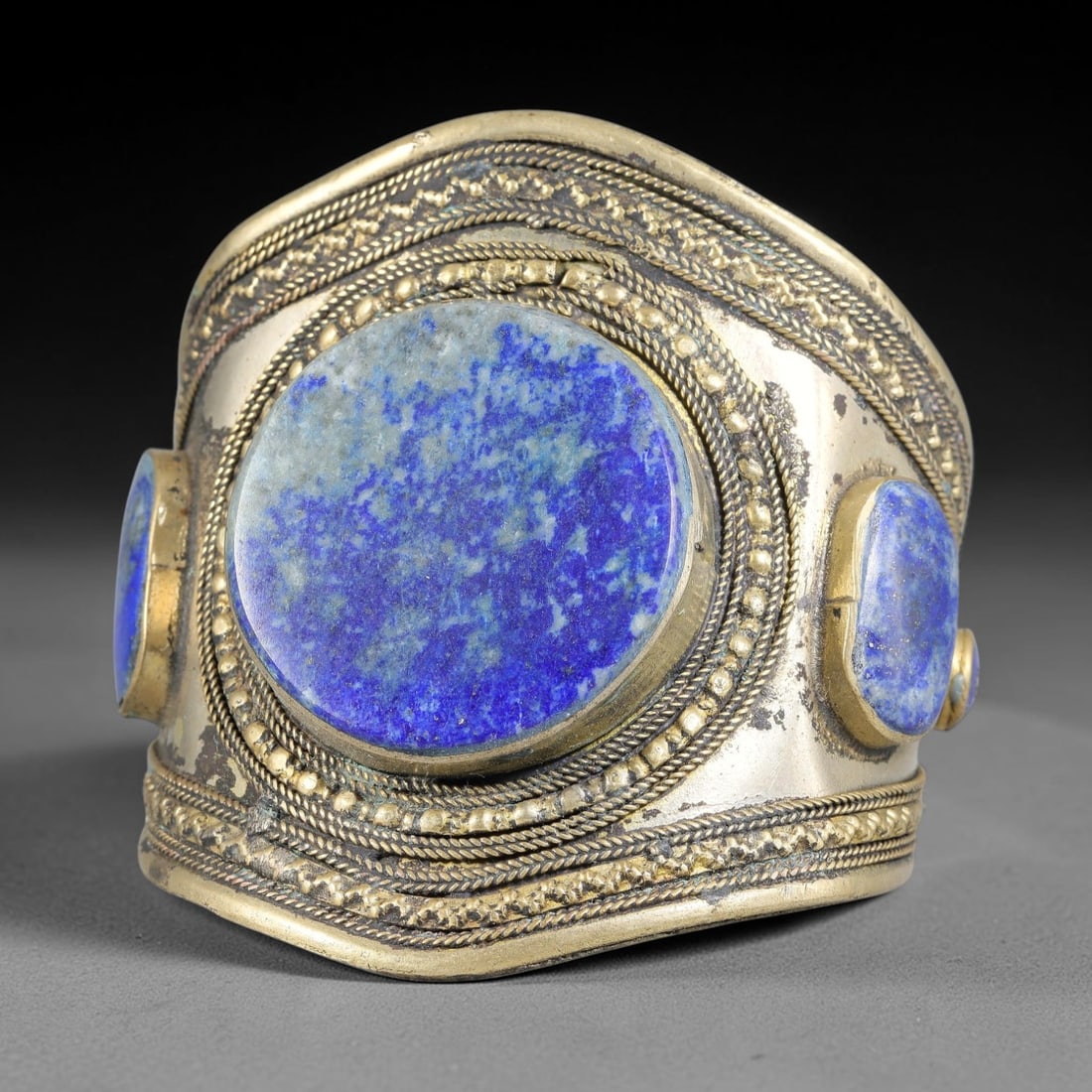A Central Asian Copper-Alloy Lapis Lazuli Inlaid Cuff Bracelet, 39AK: Wide copper-alloy cuff bracelet set with a large central circular lapis lazuli cabochon flanked by additional stone inlays, all held within heavy bezel mounts. Multiple layers of twisted filigree wire