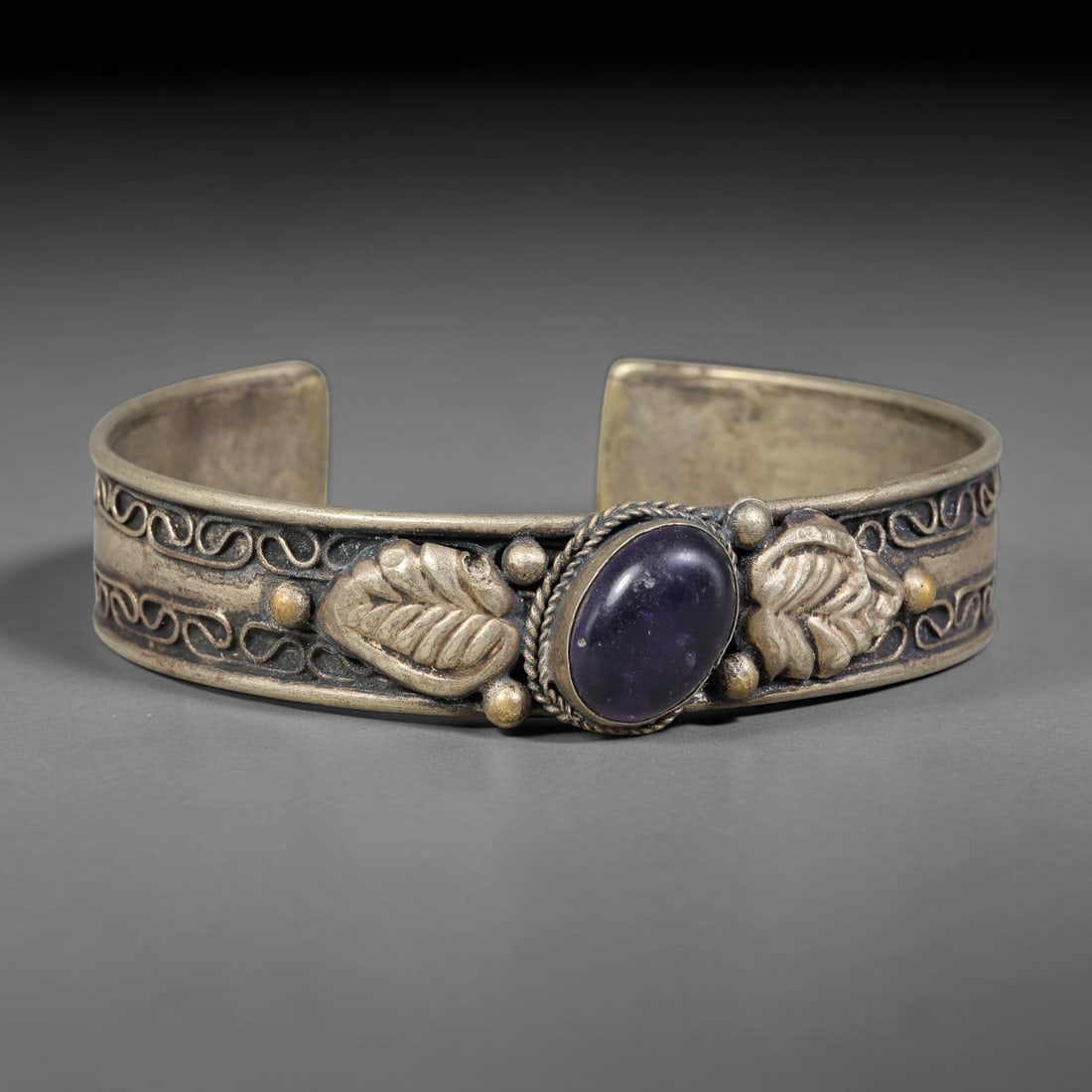 A Central Asian Copper-Alloy Amethyst Inlaid Open-Cuff Bracelet, 48AK: Open-form copper-alloy cuff bracelet set with a central oval amethyst cabochon secured within a twisted-wire bezel. Repousse leaf-shaped relief elements flank the stone, accompanied by raised spherica