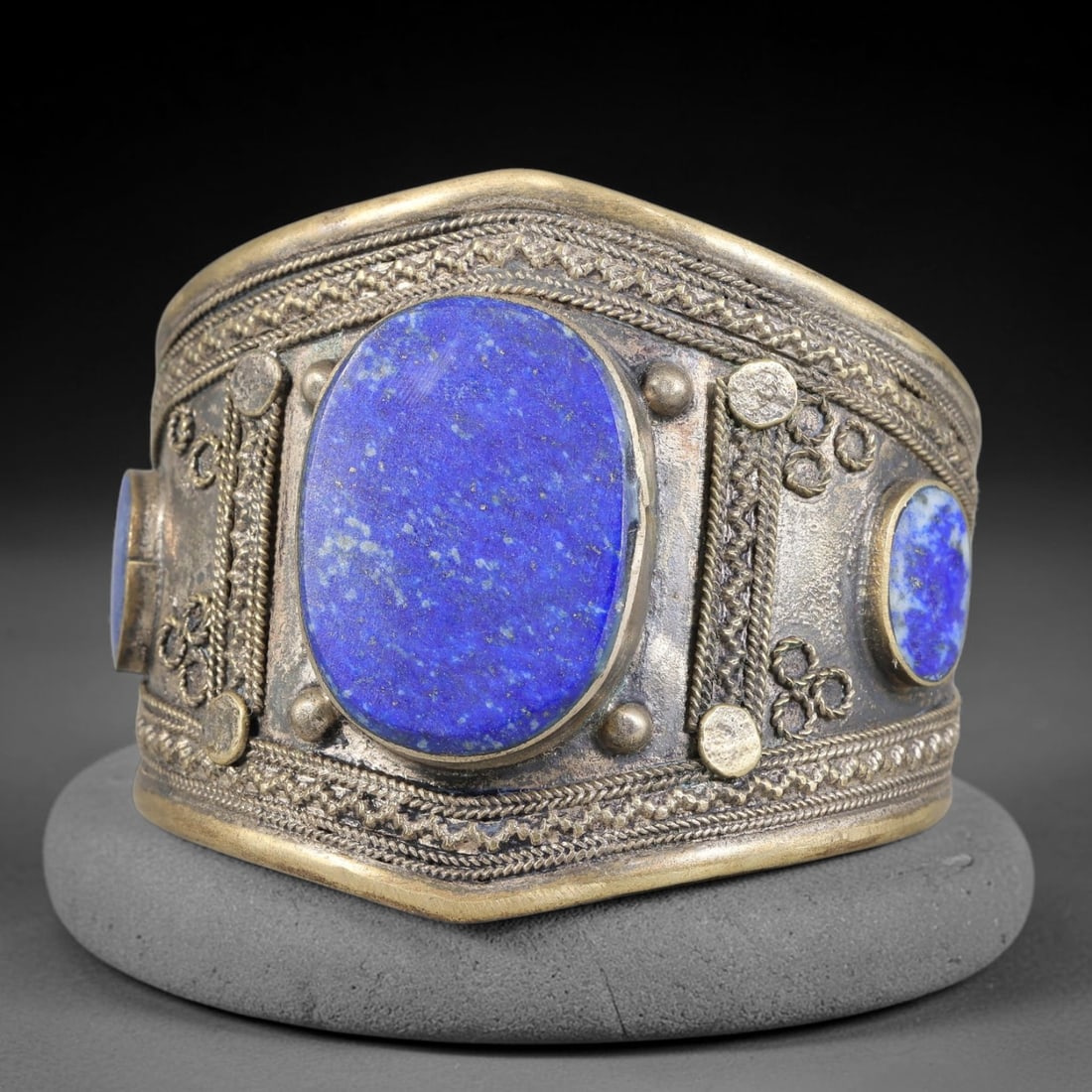 A Central Asian Copper-Alloy Lapis Lazuli Inlaid Cuff Bracelet, 43AK: Heavy constructed copper-alloy cuff bracelet set with multiple lapis lazuli oval cabochons secured in bezel mounts. The surface is enriched with applied filigree wirework, circular stamped studs and l