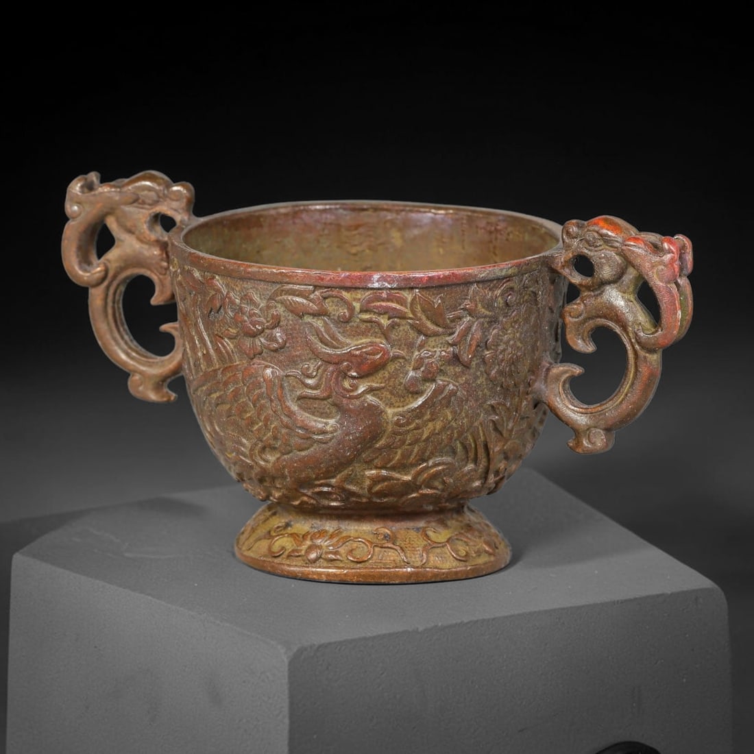 A Chinese Bronze Two-Handled Cup with Phoenix and Floral Relief Decoration, 93AD: Cast bronze cup with two open-work dragon-form handles, raised upon a decorated flared foot ring. The outer wall is richly cast in high relief with paired phoenix birds amidst scrolling peony-style fl