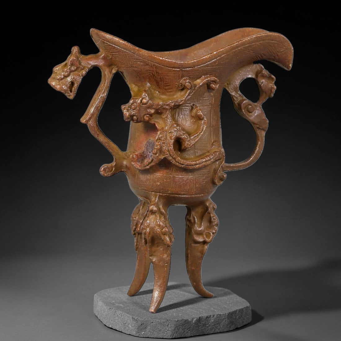 A Chinese Archaic-Style Bronze Dragon-Ornamented Jue Wine Vessel, 16AJ: Cast bronze ritual jue wine vessel with wide flared spout and elegant profile, fitted with two stylized beast-form side handles. A relief coiled dragon adorns the front body, supported by three ornate