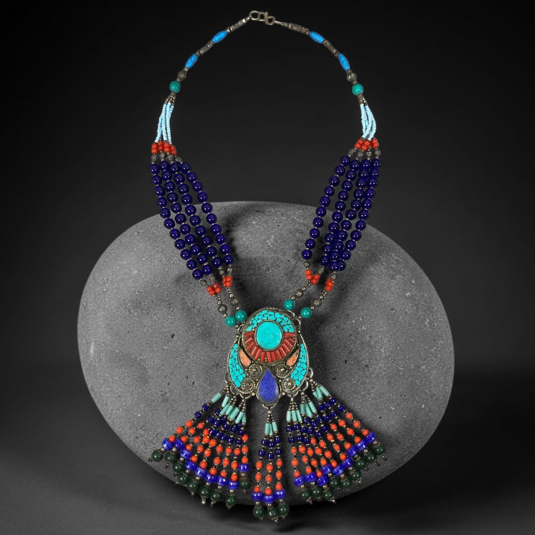 A Central Asian Multi-Strand Multi-Stone Necklace, 95AK: Multi-metal necklace composed of multiple hanging beaded strands in varied colours, anchored by an elaborate central metal pendant. The pendant features composite stone inlays secured within bezel mou