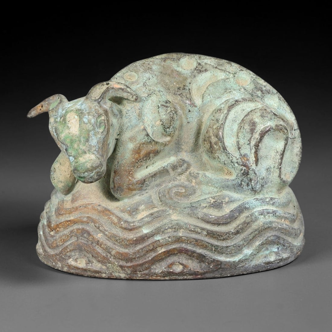 Ancient Central Asian Bronze Recumbent Bovine Effigy, 50AL: Cast bronze effigy of a recumbent bovine beast, covered in heavy mottled green and earthy brown patina with mineral encrustation across the entire surface. The animal rests with legs tucked beneath it