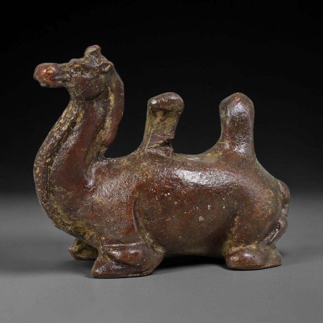 A Chinese Bronze Bactrian Camel Seated Figurine Effigy, 13AD: Cast in bronze, this figurine portrays a double-humped bactrian camel in recumbent resting pose with head raised and neck extended. Simplified robust forms capture the anatomy, with subtle textural de
