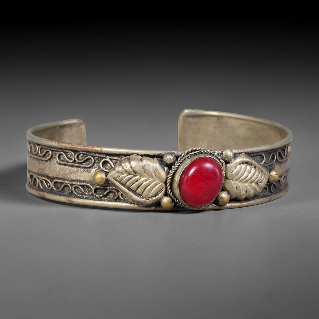 A Central Asian Copper-Alloy Red Composite Inlaid Open-Cuff Bracelet, 53AK: Open-form copper-alloy cuff bracelet centering a round red composite cabochon secured within a twisted-wire bezel. Repousse leaf-form relief ornaments flank the central stone, complemented by raised s