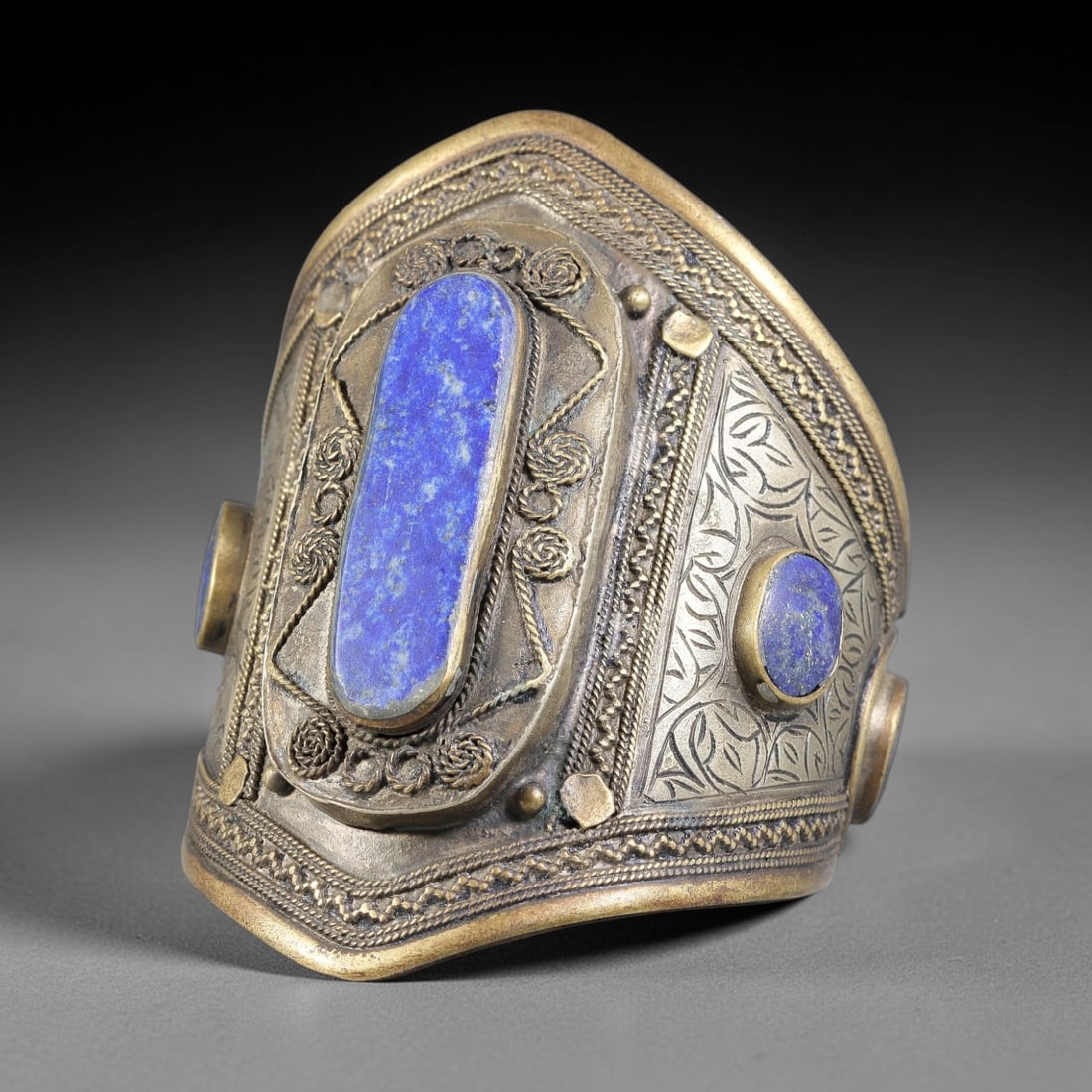A Central Asian Gilt-Copper Lapis Lazuli Inlaid Cuff Bracelet, 37AK: Broad gilt-copper cuff bracelet mounted with elongated and round lapis lazuli cabochons secured in raised bezels. The band combines applied filigree rosette motifs, twisted wire borders, and engraved 
