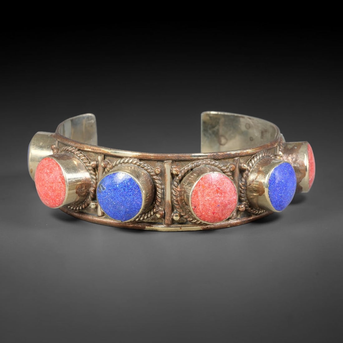 A Tibetan Copper-Alloy Multi-Stone Inlaid Open-Cuff Bracelet, 47AK: Open-form cuff bracelet worked in copper-alloy, set with circular cabochon inlays secured within raised bezel settings. Twisted cord-style wirework separates each stone mount across the band. Internal