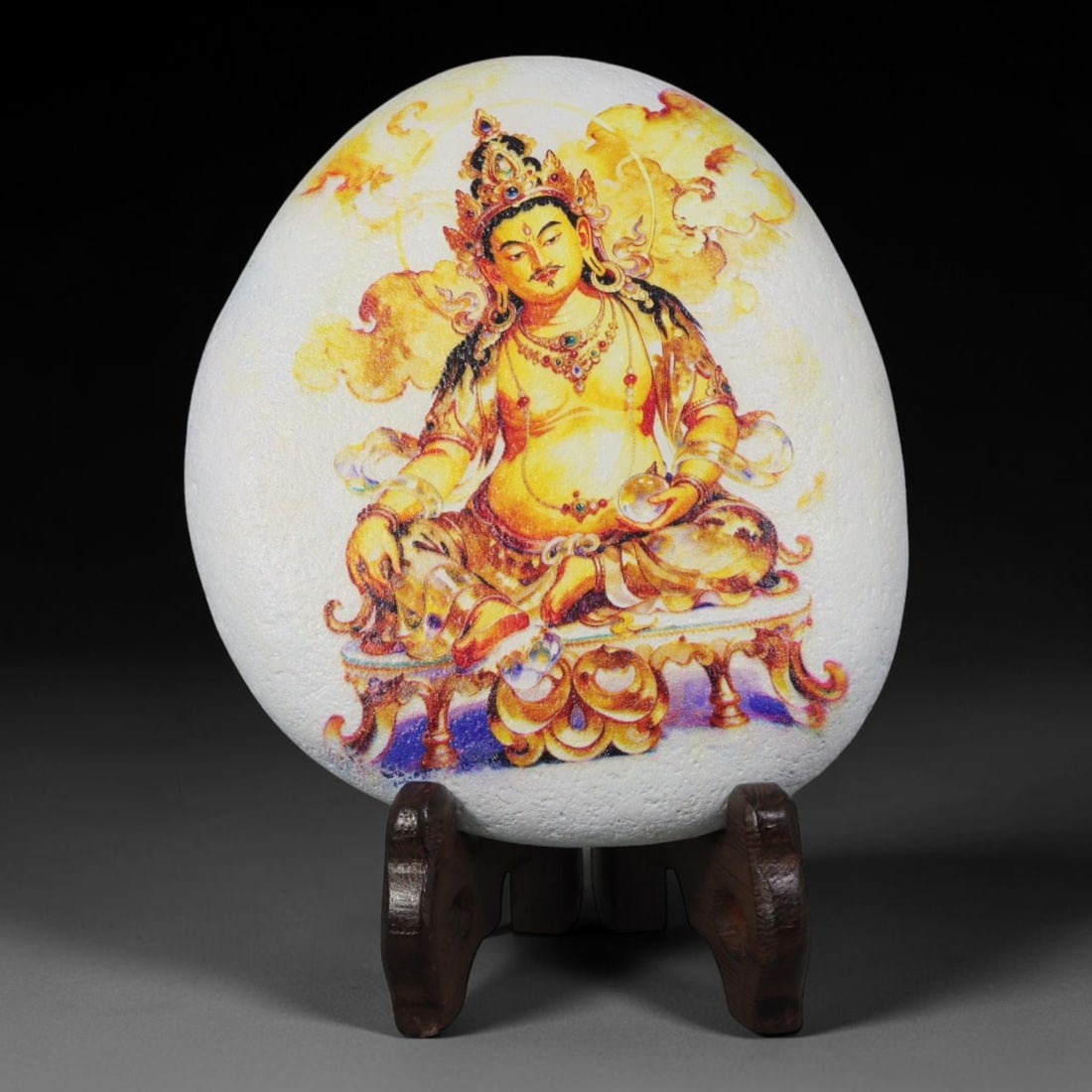 A Tibetan Thangka Style Painted River Stone with a Wealth Deity, 57BC: Painted on a naturally rounded, oval white river stone, the work depicts Yellow Jambhala, a wealth deity, in a thangka-style composition. The figure is shown in a seated posture holding a ritual imple